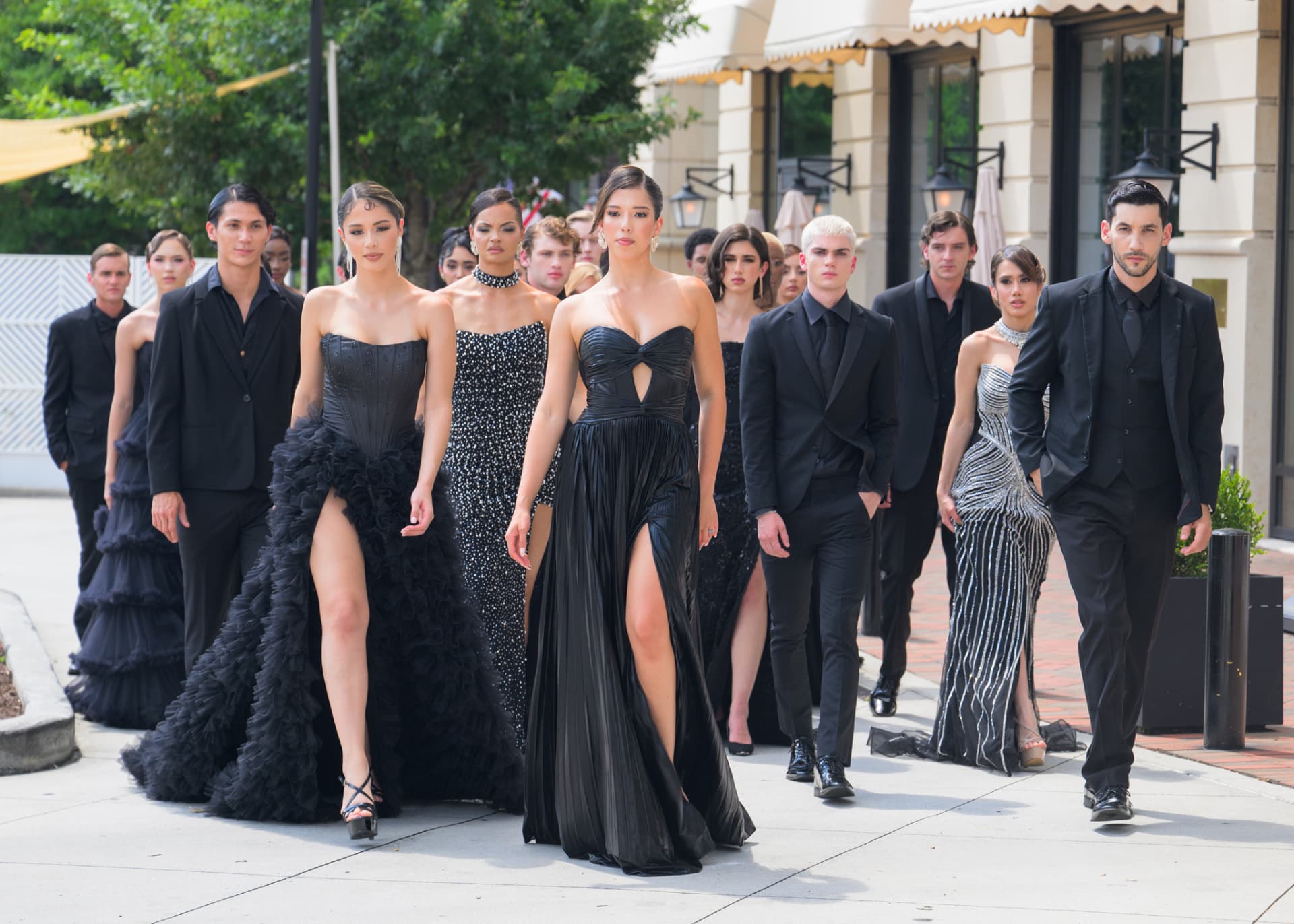 Models in black gowns and tuxedos walking a city sidewalk in formation